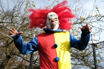 Obraz premium Halloween holiday. Scary aggressive clown with red hair jumping in a trampoline on autumn trees and blue sky background. Creepy clown costume. Festival and carnival. Horror and fear.