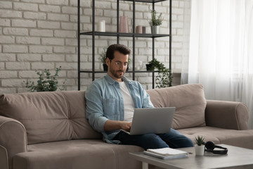 Young caucasian man in glasses sit on couch in living room work on laptop browsing internet, millennial male look at computer screen, consult client or freelance in home office, technology concept