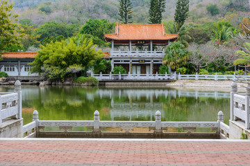 Fototapeta premium Green lake surrounded by gazebos, temples, mountains, flowers, palm grove and stone fence on the territory of Buddhist center Nanshan.