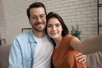 Happy young Caucasian couple hug make self-portrait picture relaxing at home together, smiling man and woman embrace take selfie on smartphone with good quality camera, lover, relations concept