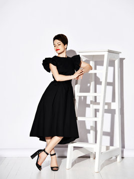 Smiling young short haired brunette woman in elegant black dress and shoes standing at leaning on big white ladder