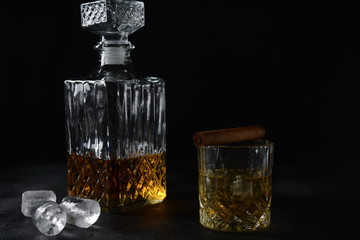 Glass of the whiskey with ice cubes and a square decanter, and cigar on a black  background.