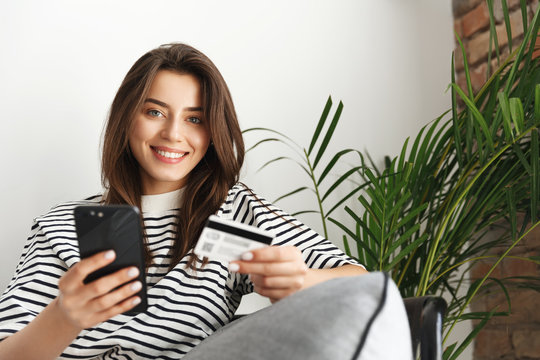 Close-up Of Relaxed Attractive Smiling Girl Sitting On Couch At Home, Checking Bank Account, Holding Credit Card And Smartphone Shopping Online For Clothes, Enter Banking Info In App, Sending Money