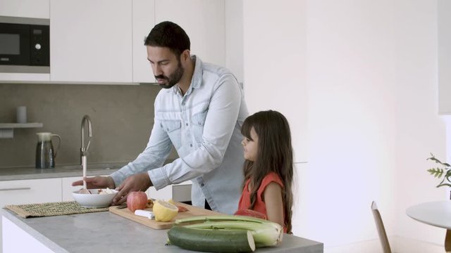 Handsome dad enjoying cooking dinner with little daughter, tossing salad, giving slice to girl to taste. Medium shot. Healthy nutrition or lifestyle concept