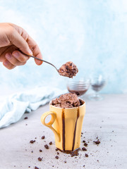 Chocolate brown ice cream in a regular yellow mug and spoon, on a blue background, with topping and crumbs. Unusual serve of dessert, copy space.