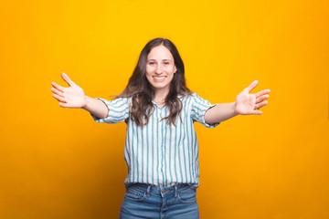 Young woman is smiling at the camera with arms wide open for hugs and embraces over yellow background.