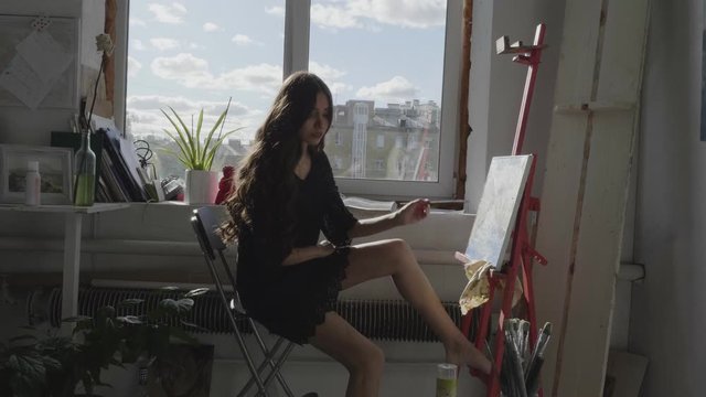 brunette girl in black dress makes smears on drawing with brush sitting near large window in art studio slow motion