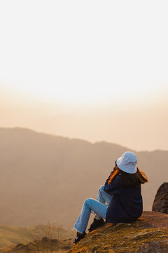 A woman with a red long-sleeved shirt and a white hat sits on the large rocks at Mulayit Taung, Myanmar. She holds a camera and looks forward to the overlapping hillsides and golden dry grasslands.