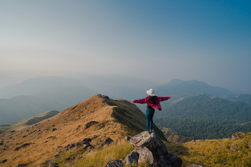 Obraz premium A woman with a red long-sleeved shirt and white hat stand and open arms on the stone at the ridge of Mulayit Taung, Myanmar. There are overlapping hills and golden dry grasslands. There is copy space.