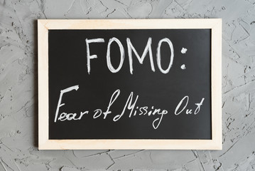 FOMO (acronym for Fear of Missing Out) concept. Handwritten text on a small blackboard with wooden frame, concrete wall as background, top view