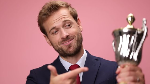 excited young businessman in navy blue suit making a hand gesture, smiling, pointing fingers to silver trophy and presenting on pink background