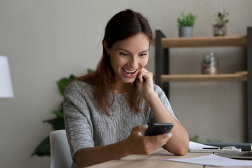 Amazed young Caucasian woman sit at desk look at smartphone screen shocked by unexpected good message, stunned happy female surprised by unbelievable sale offer or discount deal on cellphone