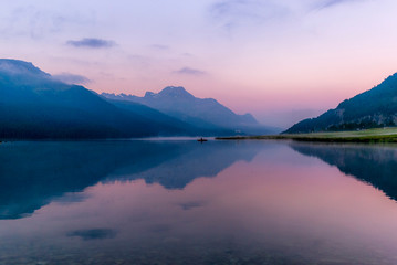 Fototapeta premium A fisherman rowing among the fog on the lake of Silvaplana in the Engadin valley at sunrise with mountains reflecting in the water
