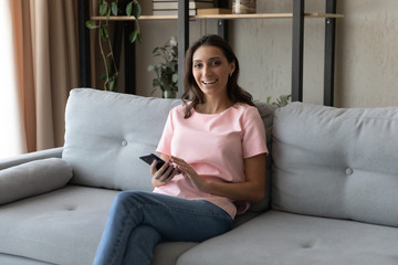 Portrait of smiling young ethnic woman sit relax on comfortable couch using modern cellphone gadget, happy arab indian female client rest on sofa text message browse interne on smartphone at home