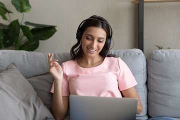 Happy young ethnic woman in headphones sit relax on couch at home talk on video call using laptop, smiling millennial mixed race female in headset speak consult client or customer online on computer