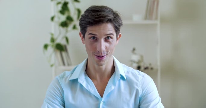 Portrait of young funny man, wears shirt sits in apartment room, looks at camera with emotional face, dances moves his head rhythmically to sides, plays with his eyebrows, indulges, flirts, close-up