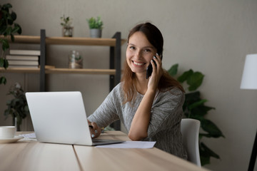 Portrait of smiling young Caucasian businesswoman sit at desk at home work on laptop talk on cellphone, happy woman have mobile online call or conversation on smartphone, technology concept