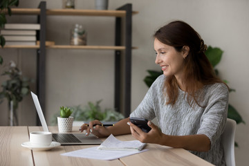 Happy young Caucasian woman sit at desk look at laptop screen use cellphone study online from home, smiling female work on computer or take web course, make notes summarize, distant education concept