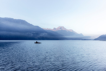 A fisherman rowing among the fog on the lake of Silvaplana in the Engadin valley at sunrise with mountains reflecting in the water