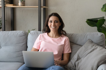 Portrait of smiling millennial Arabic indian girl sit on couch in living room use modern laptop gadget, happy young ethnic woman relax rest on sofa at home work online on computer, talk on video call