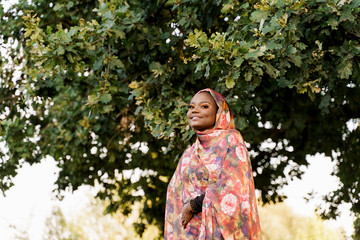 Muslim black woman African ethnicity weared traditional colorful hijab smiles under green tree. Arabic islam religion clothes. Pretty black girl smiles. Business for black woman.