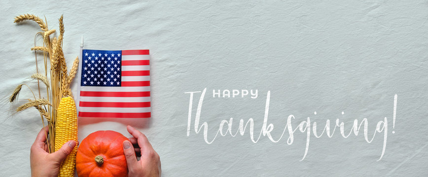 Happy Thanksgiving! USA American flag on white textile, hand holding corn cob and wheat ears, orange pumpkin. Banner design, panoramic image with text.