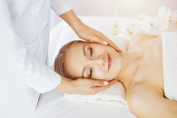 Fototapeta na wymiar Beautiful caucasian woman enjoying facial massage with closed eyes in sunny spa salon. Relaxing treatment in medicine and Beauty concept