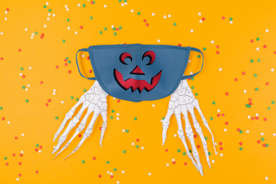 Reusable blue anti-virus mask with Jack lantern print and paper skeleton hands. Orange background with decorative stars. Flat lay. The concept of protection from the virus during Halloween