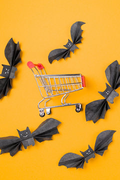 The customer's toy cart is located in the middle of paper decorative bats. Orange background. Vertical. Flat lay. The concept of Halloween and holiday shopping
