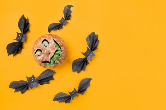 A pumpkin with an image of a face surrounded by bats made of paper. Orange background. Flat lay. Copy space. Concept of Halloween and holiday decorations