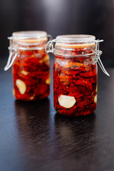 Sun-dried tomatoes are packed in a jar with spices and garlic and drenched in olive oil. Tins of tomatoes are on a black table, with slices of tomato and spices scattered nearby.