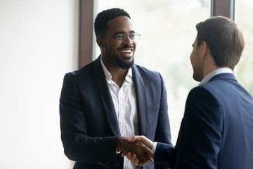 Satisfied african insurer, manager or consultant shaking hand to european customer, happy diverse male business partners completing a deal with handshaking, two colleagues employees meeting in office - Powered by Adobe