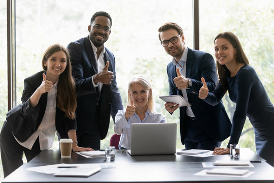 Happy senior woman team leader and diverse millenial employees looking at camera showing thumbs up, group of young workers or consultants headed by older boss recommend their service. Portrait