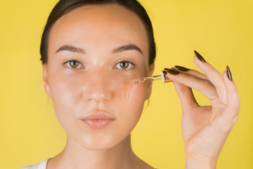 Fototapeta premium A beautiful young girl applies a serum to her face with a pipette. Cosmetology, skin care. Yellow background