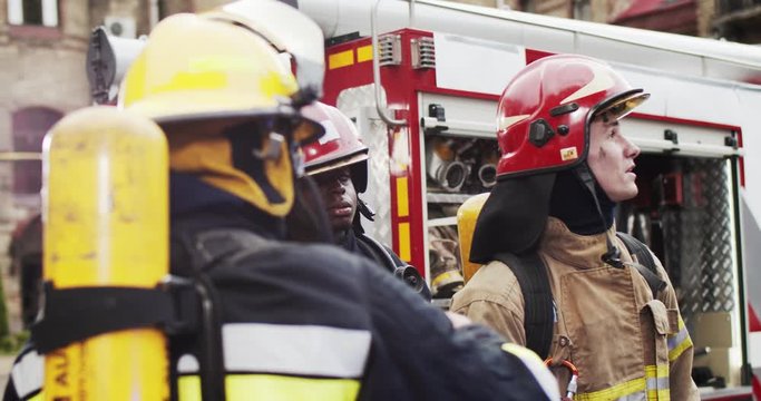 Three firefighters of different races in helmets and gull equipment discussing rescue plan standing near fire van. Firefighter with tablet in hands showing direction. Smoke from fire covers rescuers.