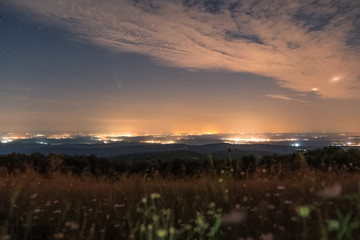 Comet Neowis in the night sky with clouds, cities shining in the background, view of the landscape from the hill
