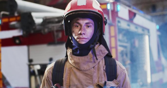 Portrait of firefighter approaches from fire van with flashing lights on and standing in front of camera looking after hard work. Smoke from fire covers rescuers. Concept of saving lives, fire safety