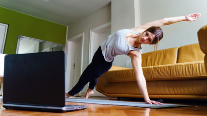 Young caucasian woman practicing yoga at home beautiful instructor teaching online class training in apartment standing in position asana in front of laptop using video in day or morning - new normal