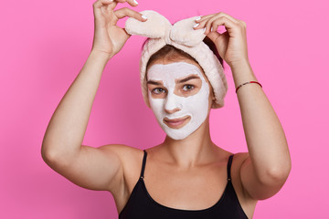Beautiful woman with white clay facial mask on face, touching her hairband and looking directly at camera, beauty posing isolated over rosy background.