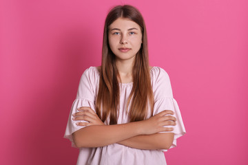 Fototapeta premium Portrait of serious young woman with pleasant appearance wearing pale rose posing at pink wall with arms crossed, lady in dress, looking at camera.