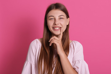 Attractive woman with happy look, standing in fashionable outfit, smiling while holding index finger on lip, expressing positive emotions, being curious over pink background.