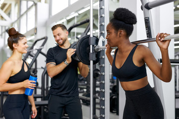 two athletic diverse women talking with trainer in the gym, they communicate, discuss something, smile. healthy lifestyle concept