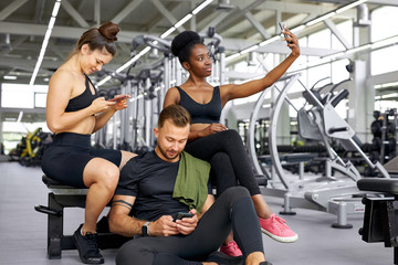 Fototapeta na wymiar portrait of tired diverse fit people enjoying time after sport exercises, african and caucasian team sit together in gym, using smartphone