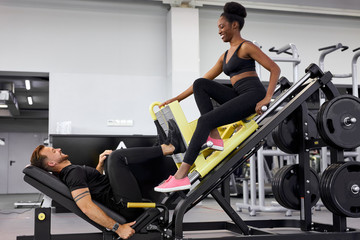 Fototapeta na wymiar athletic people support each other in gym, do exercises together. man pumping legs on equipment with weight, african woman sit near, smile