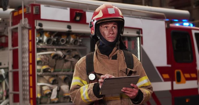 Portrait of fireman in costume and helmet tappig on tablet computer and looking on sides. Smoke from fire covers rescuers. The concept of saving lives, heroic profession, fire safety