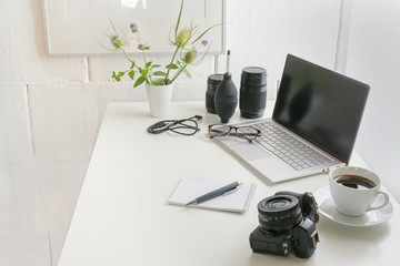 White office desk with laptop, camera, lenses, cable, glasses, notepad and a coffee cup, modern workplace of a photographer against a bright wall, copy space, selected focus