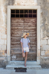 Fototapeta na wymiar Beautiful young female tourist woman standing in front of vinatage wooden door and textured stone wall at old Mediterranean town, smiling, holding, smart phone to network on vacationes.