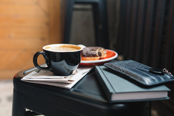 Close up of cup of coffee with milk, donut, newspaper, wallet, notebook on black table. Still life, lifestyle concept. Stylish composition.
