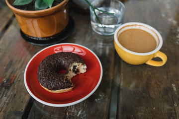 Chocolate bitten-into donut on plate, cup of coffee and glass of water on background.