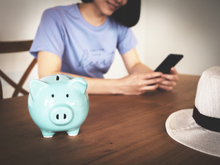 piggy bank on wooden table with blurry background of woman using smart phone, booking hotel online, saving money for vacation concept, lifestyle saving, with copy-space